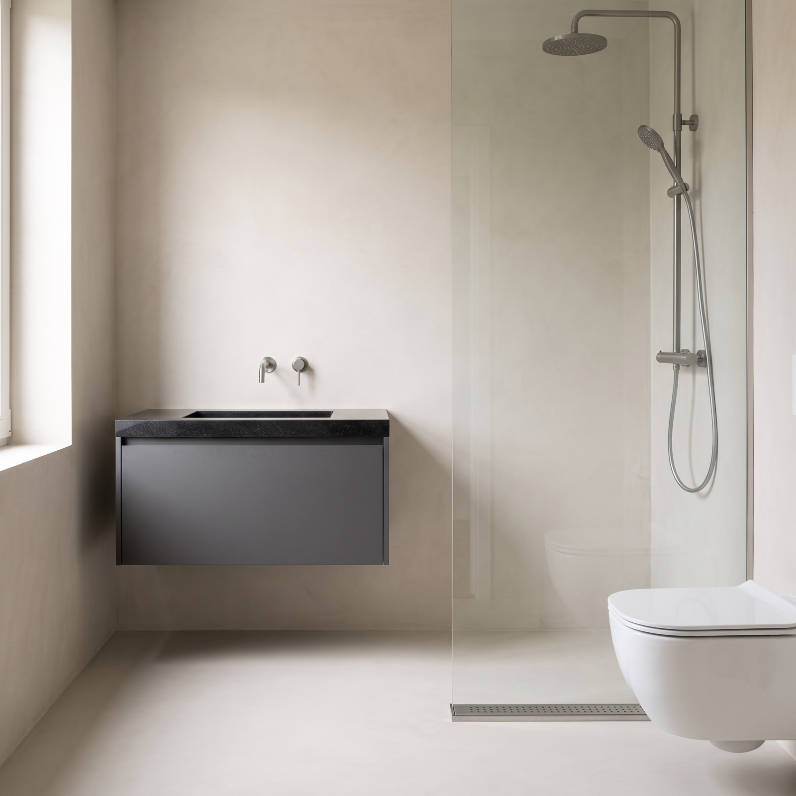 Badkamer met een antraciet badmeubel met een natuurstenen wastafel, een doucheset in gunmetal op de wand, een douchegoot in de vloer en een wit wandcloset