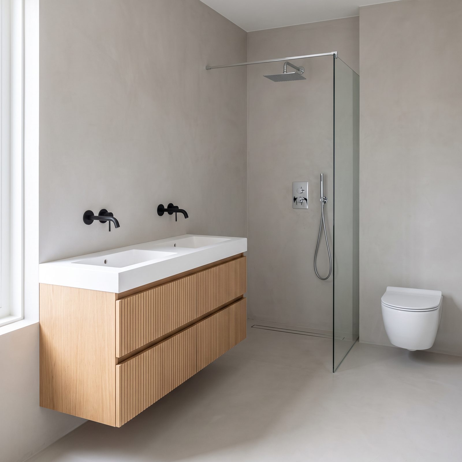 Badkamer met een licht eiken badmeubel met ribbelfronten en een dubbele witte wastafel, een chromen inbouwregendouche achter een glazen wand, een douchegoot in de vloer en een wit wandcloset
