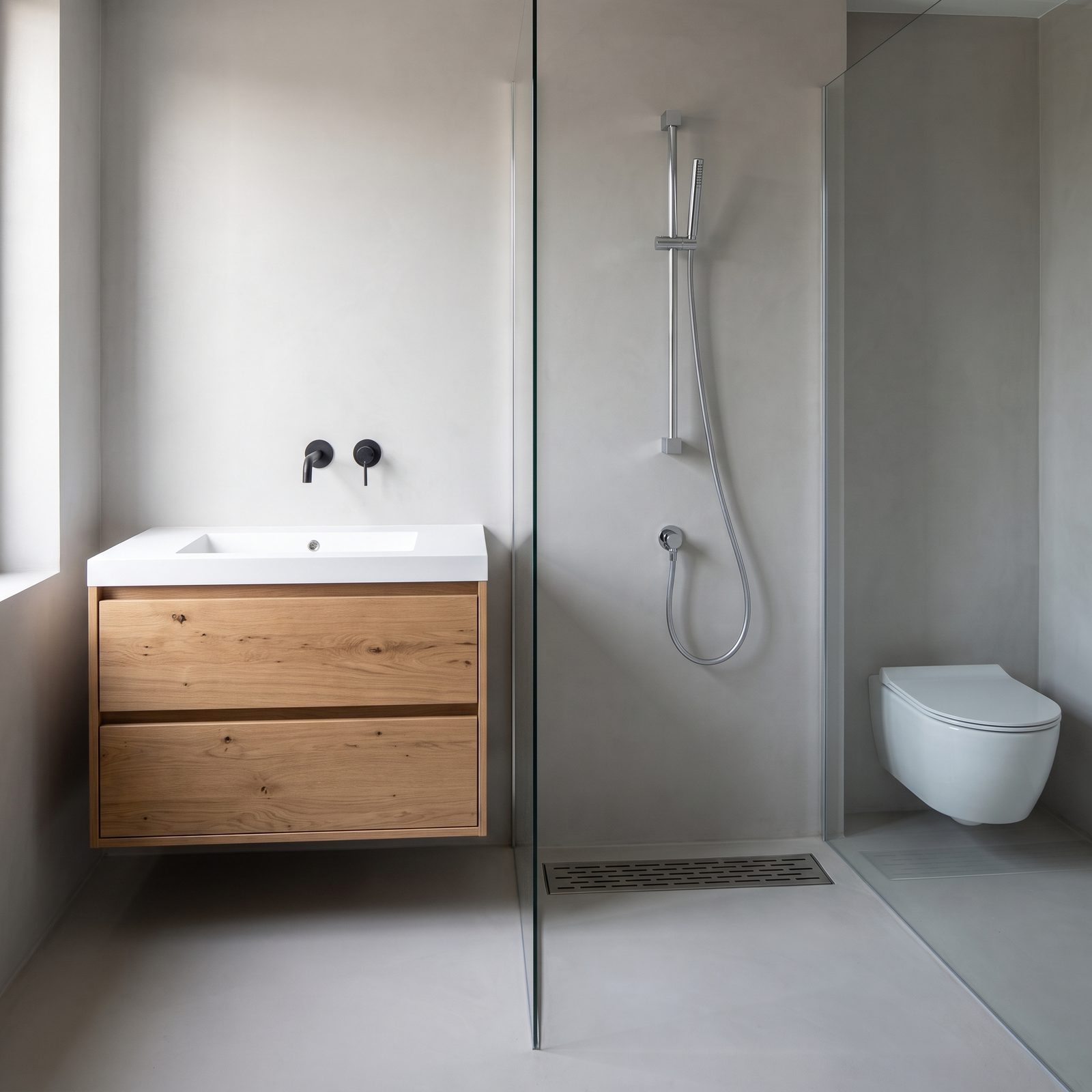 Badkamer met een warm eiken badmeubel met een witte wastafelplaat en een mat zwarte wandkraan, een chromen glijstangset met handdouche achter een glazen wand, een douchegoot in de vloer en een wit wandcloset